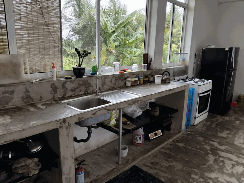 Villa Idda's private kitchen with a concrete worktop, sink, gas cooker and fridge, looking out onto banana trees